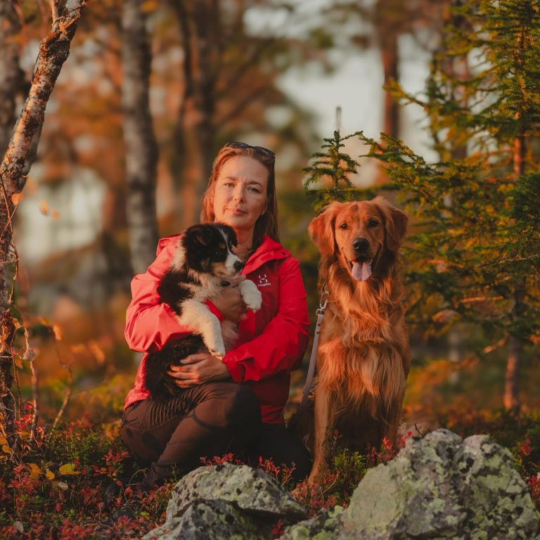 Sini Lindroosin kuva. Hän on kuvassa syksyisessä metsässä istumassa kahden koiransa kanssa.